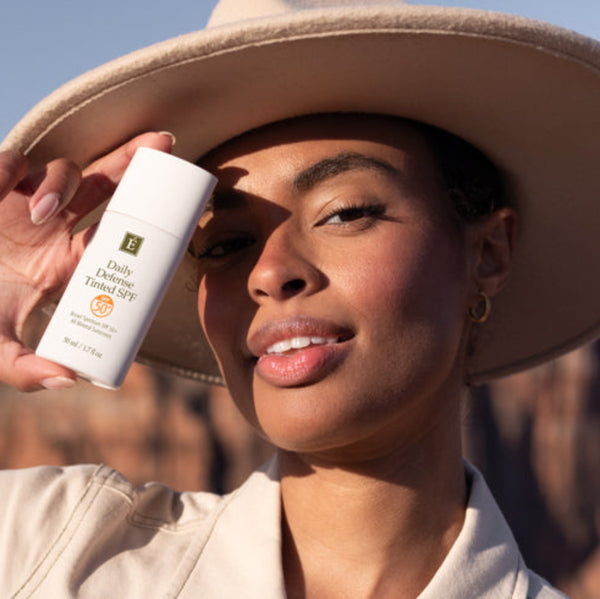 A woman wearing a beige wide-brim hat and matching top holds a white bottle of JUVENIAL Daily Sun Protection SPF 50+ in front of her face, with a desert canyon landscape in the background.