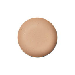 A circular makeup swatch showing a warm, light tan skin tone with subtle speckles, centered on a solid white background.