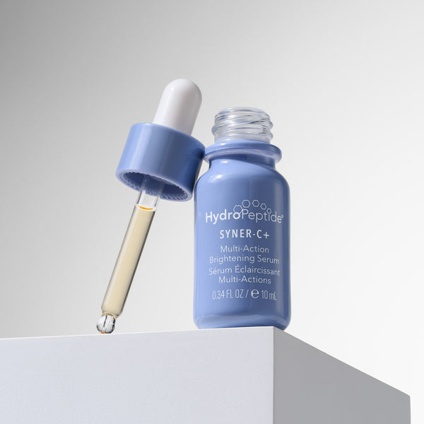 A small blue glass serum bottle labeled HydroPeptide SYNER+C with a dropper cap resting beside it on a white pedestal against a light gray studio background.