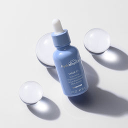 A light blue dropper bottle labeled Hydrology Syringe C+ on a clean white background, surrounded by four translucent spherical props casting soft shadows.