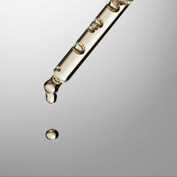 A close-up of a glass dropper containing a transparent golden liquid, with one droplet falling below and a blue cap visible at the top against a light gray gradient background.