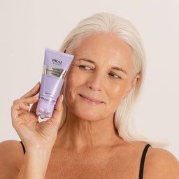 A woman with platinum blonde hair holds up a lavender tube of PRAM hair growth serum against a plain off-white background.