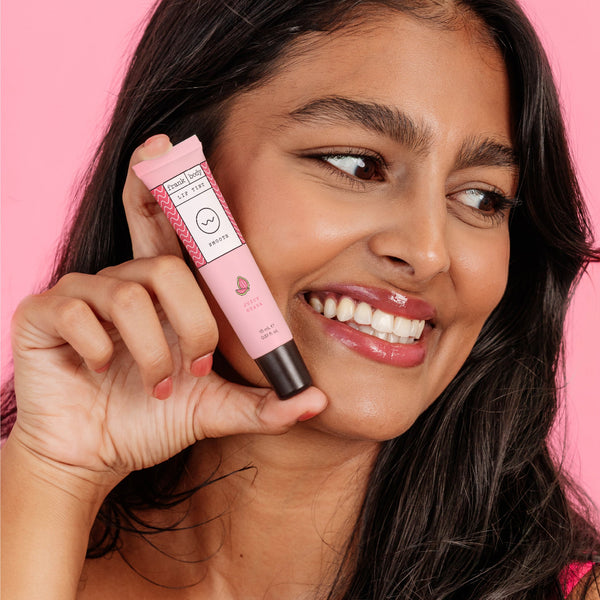 A woman with long dark hair holds a pink tube of lip balm against a solid pink background. The tube is light pink with a black cap and white text, and the woman is smiling and looking to the side.