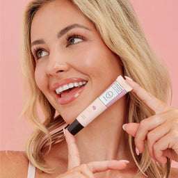 A smiling blonde woman holds a tube of makeup near her neck against a soft pink background. The tube is pale pink with a black cap and a label that reads 'The Beauty Brand' and 'Soft Pinch'.