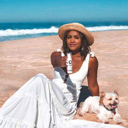 a woman holding a bottle of PRIORI TETRA Broad Spectrum SPF 55 with Universal Skin Tint while sat on a beach with a dog