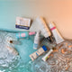 An overhead flat-lay of multiple skincare and beauty products arranged on a translucent aqua tray over rippling water. Visible products include Dr.