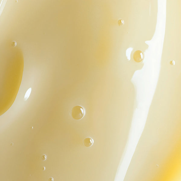 A close-up texture shot of a smooth, glossy cream-colored cosmetic gel or liquid with small air bubbles, showing a warm pale yellow hue and soft highlights.