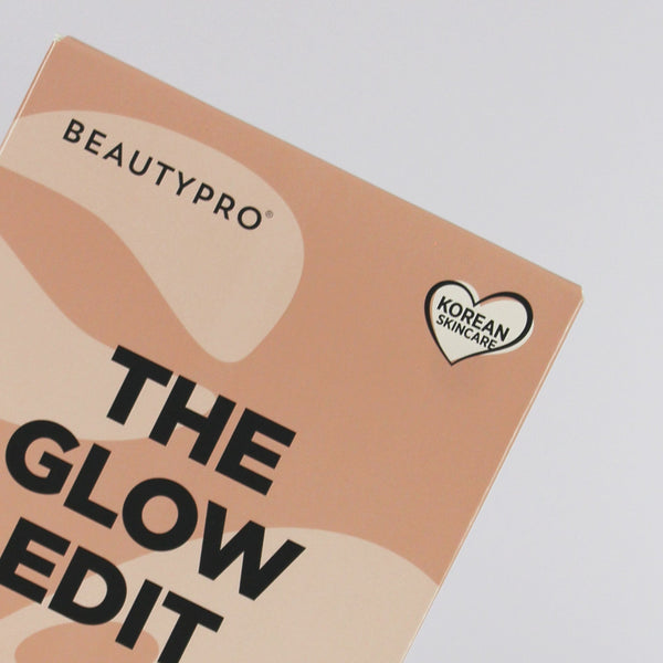 Close-up of a Beautypro cosmetic box titled 'The Glow Edit' with abstract peach and beige shapes, bold black text, and a small heart-shaped Korean Skincare badge on a light gray background.