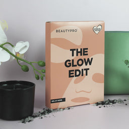 A BeautyPro 'The Glow Edit' spa at home kit box, styled on a light surface with a black candle, white orchid flowers, and a green pouch in the background.