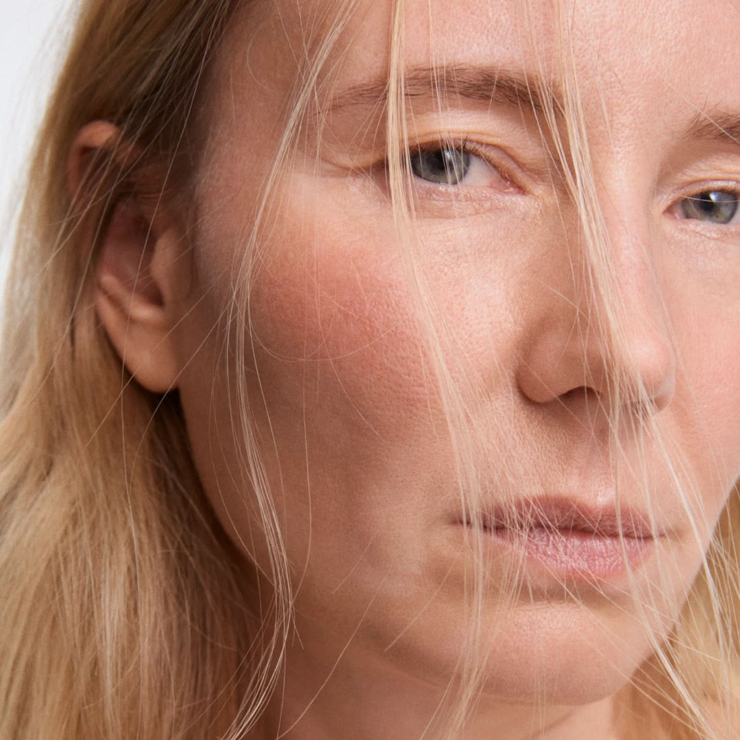 Close-up portrait of a woman with blonde hair partially covering her face, shown against a plain light background.