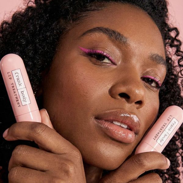 A close-up lifestyle shot of a woman holding two pink lip balm tubes labeled 'famously good' against a solid pink background.