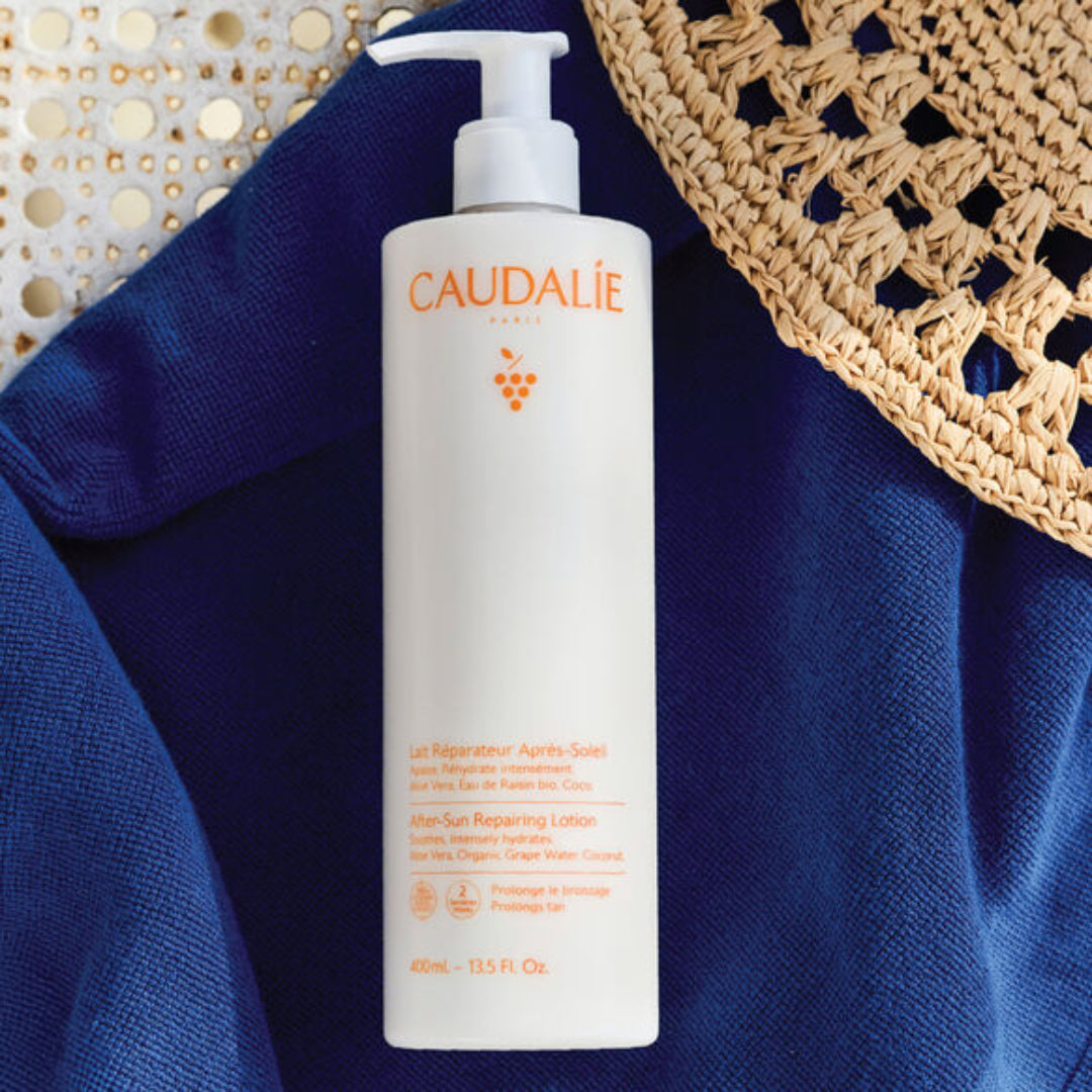 Caudalie after-sun repairing lotion bottle on a blue fabric background with a woven bag.