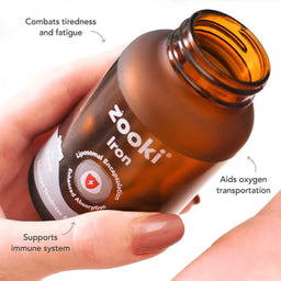 A hand holding an open amber brown bottle labeled 'Zooki Iron' with text callouts describing benefits: 'Combats tiredness and fatigue,' 'Aids oxygen transportation,' and 'Supports immune system.'.