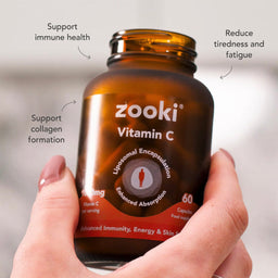 A hand holding an amber brown bottle of zooki Vitamin C capsules, with text callouts around it highlighting benefits like support immune health, reduce tiredness and fatigue, and support collagen formation.