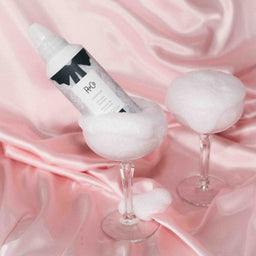 a can of R+Co Chiffon Styling Mousse in a Champaign glass willed with foam