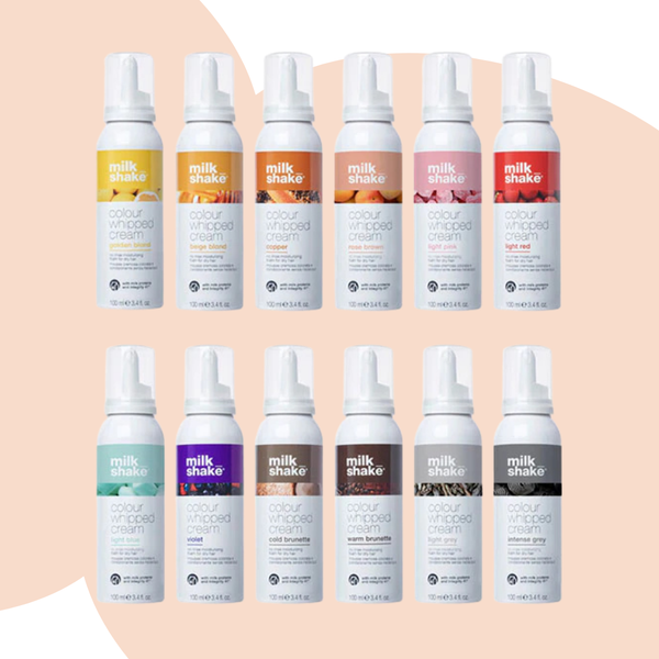 Twelve Milk Shake Colour Whipped Cream spray bottles arranged in two rows on a soft peach background. The top row features warm-toned variants in yellow, orange, peach, pink, and red, while the bottom row shows cooler-toned variants in mint, purple, brown, black, and gray.