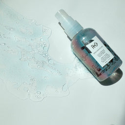 a closeup of a R+Co DREAMHOUSE Cold Pressed Watermelon Wave Spray bottle with its contents spilled out