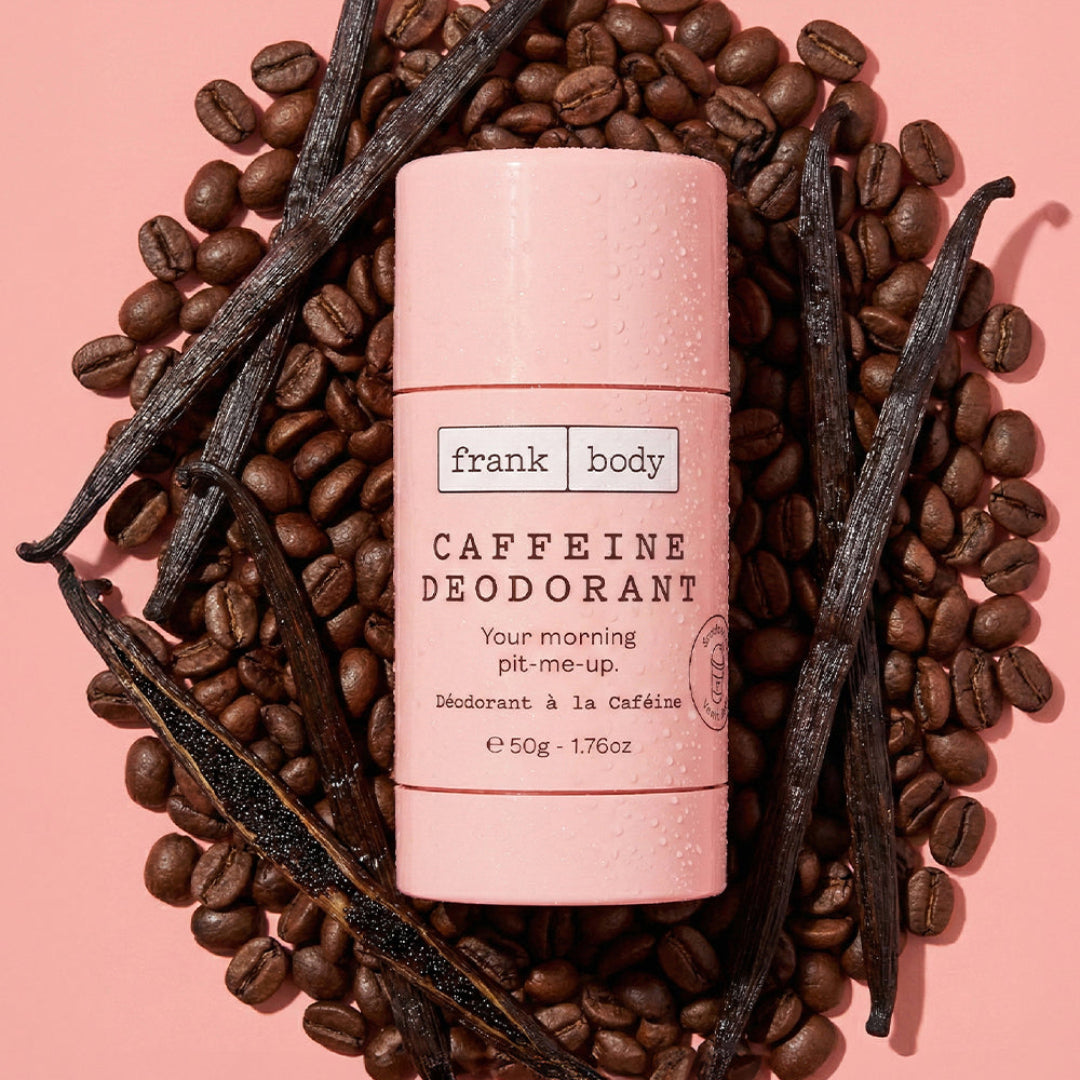 A pink Frank Body caffeine deodorant stick sits in the center, surrounded by scattered coffee beans and vanilla beans on a soft pink background.