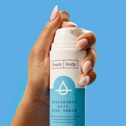 Hand holding a 'frank body' hyaluronic acid body serum against a blue background