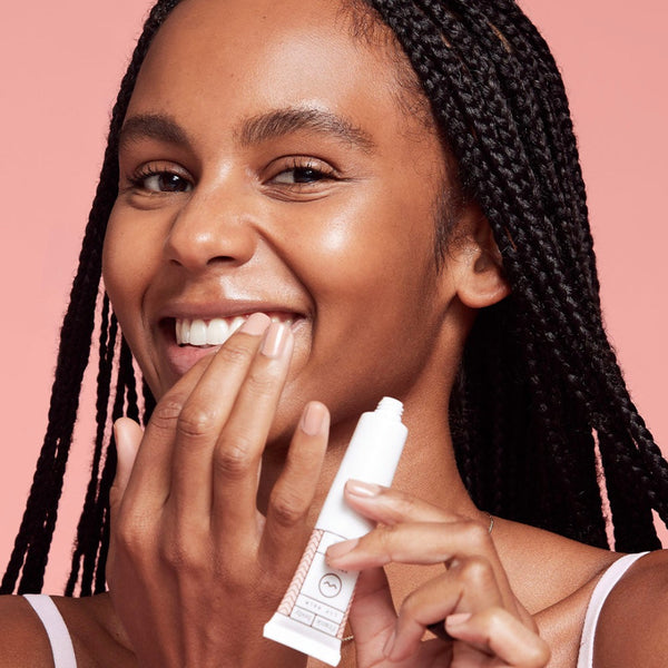 Woman applying a white tube of lip balm to her lips against a pink background