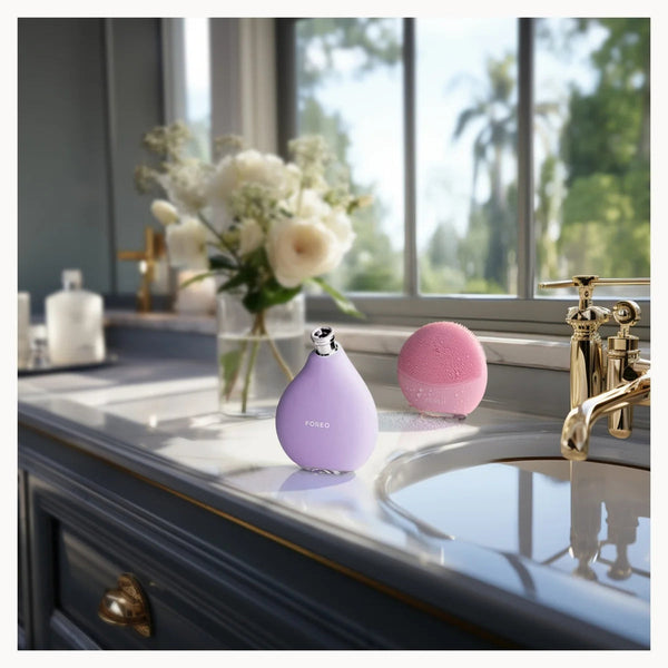 Two small personal care devices, one lavender and one pink, displayed on a bathroom counter next to a sink and faucet, with a vase of white flowers in the background.