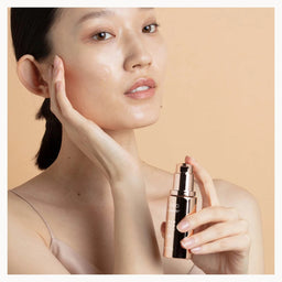 A woman with clear, radiant skin holds a small black and rose gold cosmetic pump bottle against a soft beige background.