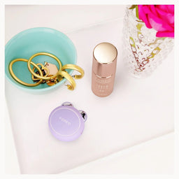 A small round purple and silver key finder with the brand name 'FONKO' printed on the lid, styled on a white tray alongside a mint bowl with gold rings, a rose gold bottle, and a pink flower in a glass vase.