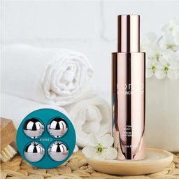 A rose gold Foego Skin Boost bottle standing on a small wooden tray next to a teal circular case holding four silver reflective spheres, styled with white towels, flowers, and a woven mat in a bright spa-like setting.