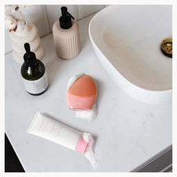 A makeup blending sponge with pink and peach sponge heads resting on a white marble bathroom counter next to a white sink, with a tube of cream and other toiletries nearby.
