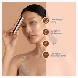 A woman holds a cylindrical cosmetic tube against her cheek on a warm beige background, with text callouts on the right stating dermatologist tested, suitable for all skin types, vegan, and 91% natural origin ingredients.