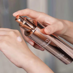 A close-up of a person's hands holding a metallic rose gold cosmetic bottle and spraying a clear liquid onto the back of another hand.