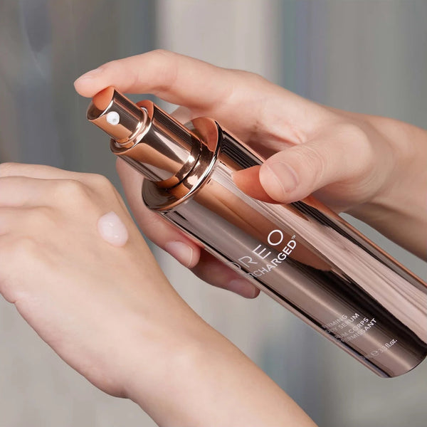 A close-up of a person's hands holding a metallic rose gold cosmetic bottle and spraying a clear liquid onto the back of another hand.