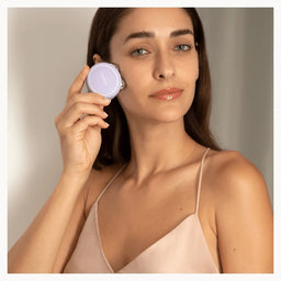 A woman with long brown hair holds a round lavender compact mirror near her face, wearing a light beige spaghetti-strap camisole against a neutral gray background.