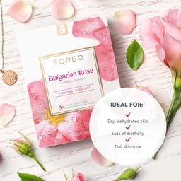 FOREO Bulgarian Rose sheet mask packaging shown on a light wood surface with pink rose petals, buds, and leaves. A white circular badge lists ideal skin conditions, and a gold pendant necklace is visible at the upper left.