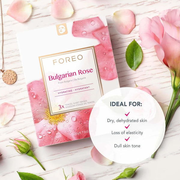 FOREO Bulgarian Rose sheet mask packaging shown on a light wood surface with pink rose petals, buds, and leaves. A white circular badge lists ideal skin conditions, and a gold pendant necklace is visible at the upper left.