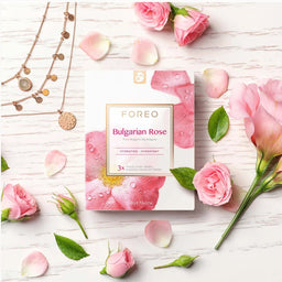 FOREO Bulgarian Rose skincare packaging box styled in a flat lay with pink roses, petals, and gold necklaces on a white wood background.