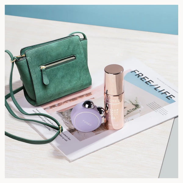 A green suede crossbody handbag with a zippered front pocket and matching strap, styled on a magazine page next to a rose gold cosmetic bottle and a small lavender round pouch.