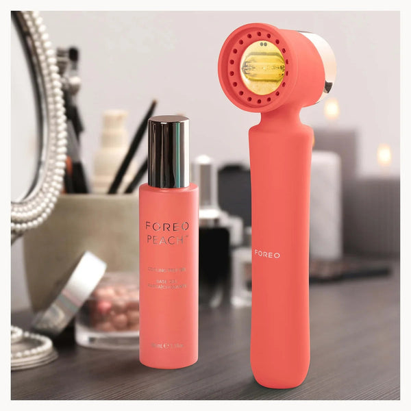 A FOREO facial cleansing device in coral pink stands next to a matching FOREO bottle with a silver cap on a dark tabletop, with blurred cosmetic items and a mirror in the background.
