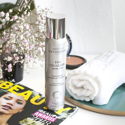 Institut Esthederm Cellular Water Mist on a night stand surrounded by flowers and magazines 