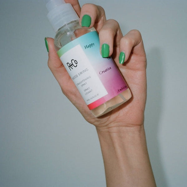 a hand holding a bottle of R+Co Mood Swing Straightening Spray to the air