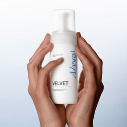 A person's hands holding a white pump bottle of Avval Velvet hand cream against a light blue gradient background.