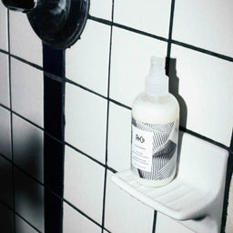 a bottle of R+Co Pinstripe Intense Detangling Spray next to a shower