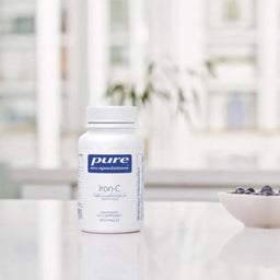 A white bottle of Pure Encapsulations Iron-C dietary supplement placed on a reflective white surface, with a softly blurred kitchen background and a small bowl of blueberries to the right.