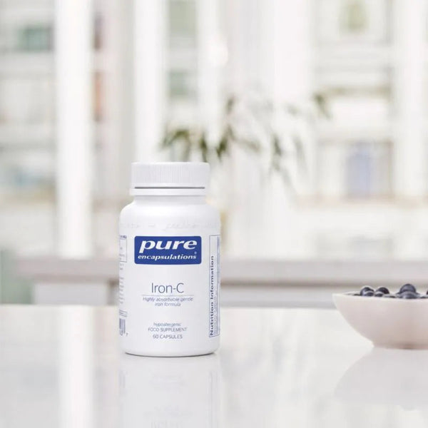 A white bottle of Pure Encapsulations Iron-C dietary supplement placed on a reflective white surface, with a softly blurred kitchen background and a small bowl of blueberries to the right.