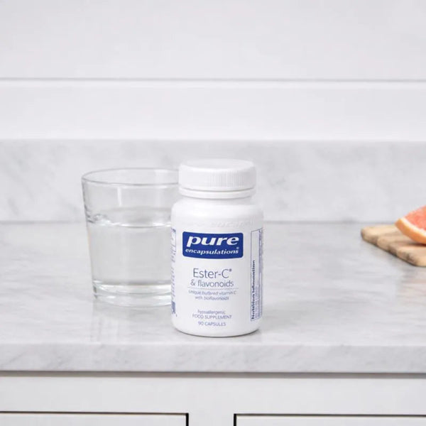 A white bottle of Pure Encapsulations Ester-C & Flavonoids dietary supplement stands on a light marble countertop next to a clear glass of water, with a blurred citrus slice in the background.