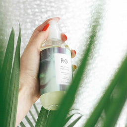 a bottle of R+Co Relative Paradise Fragrance Spray being sprayed in front of a plant