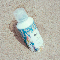 a bottle of R+Co Sail Soft Wave Spray sat in the sand