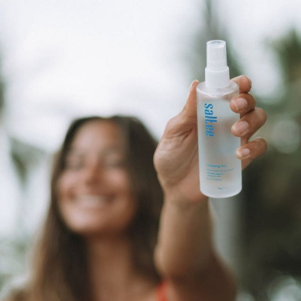 a woman out of focus holding a bottle of Saltee Hydrating Mist to the camera