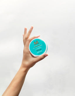 a hand holding a tub of Moroccanoil Texture Clay