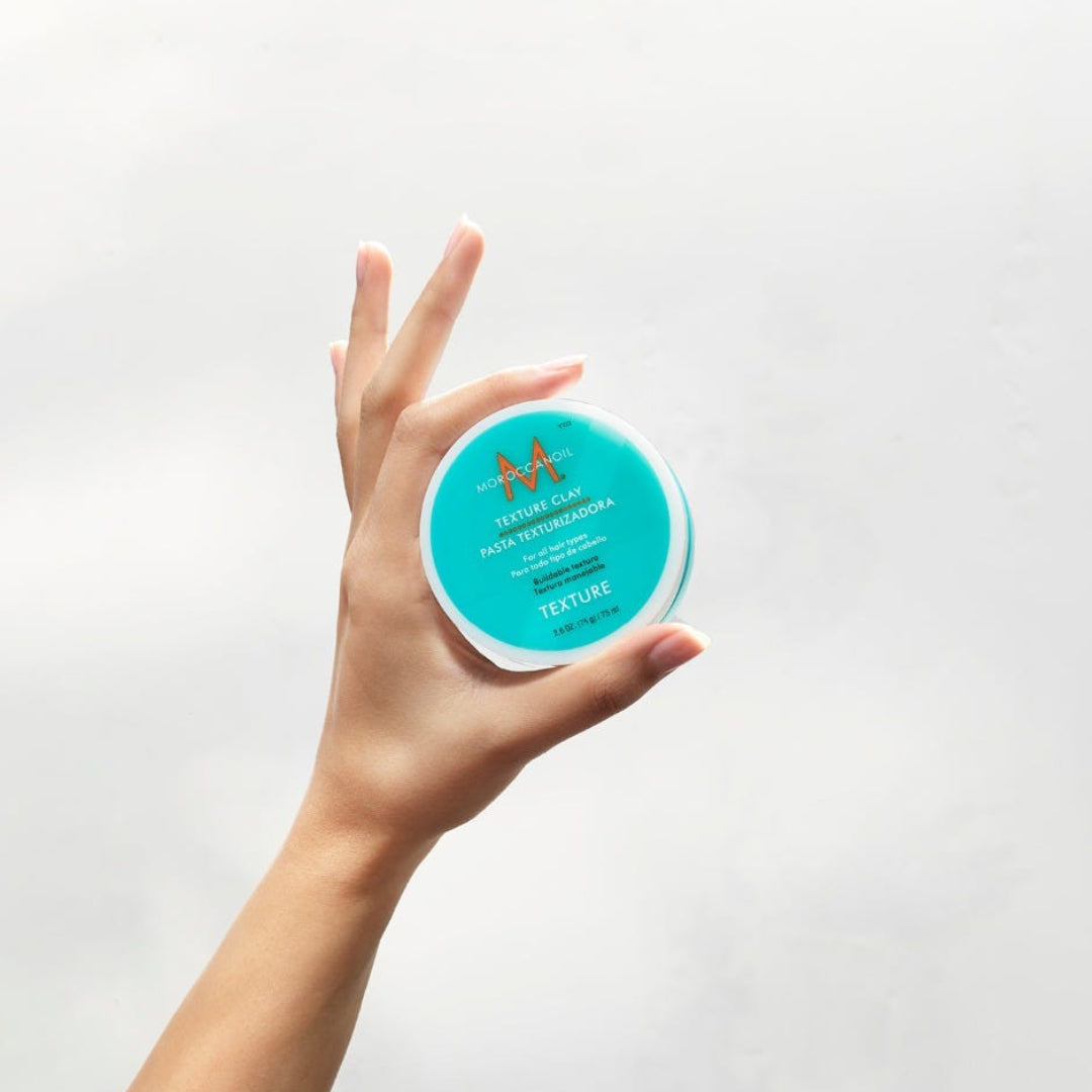 a hand holding a tub of Moroccanoil Texture Clay